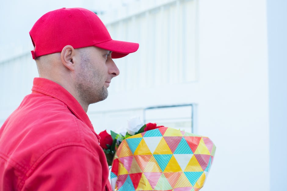 A man wearing a red baseball cap and red jacket is holding a floral gift wrapped in vibrant, geometric-patterned paper with triangles in shades of yellow, blue, red, and orange. The bouquet appears to