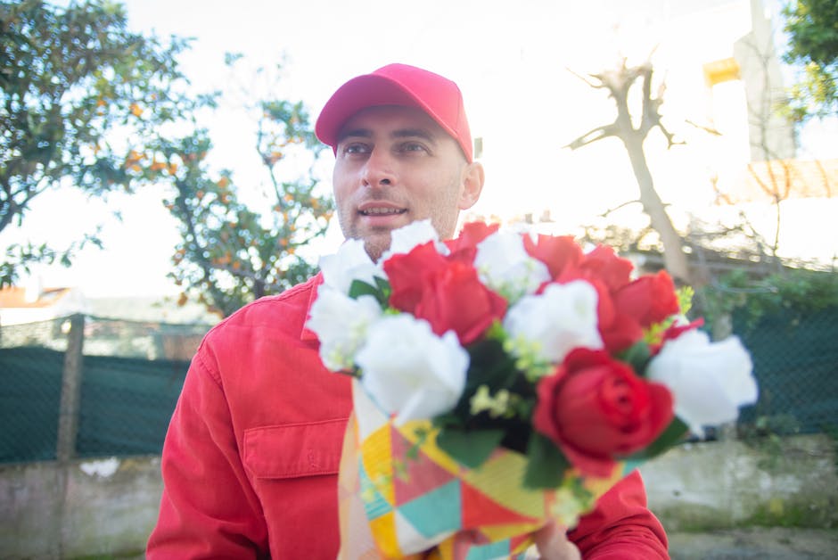 A male delivery person dressed in a red jacket and matching cap holds a vibrant bouquet of fresh flowers, including red, white, and green blossoms, wrapped in colorful patterned paper. He is outdoors,