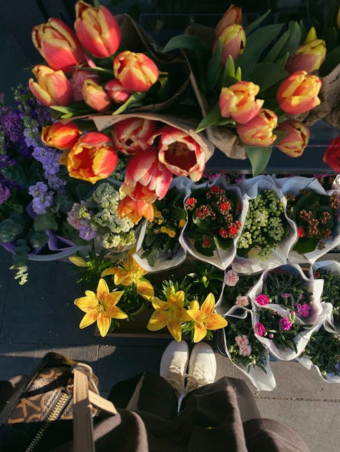 A top-down view of a selection of fresh floral arrangements and bouquets displayed on a dark surface at floristforestgate.co.uk. The central bouquet features vibrant orange and yellow tulips with gree