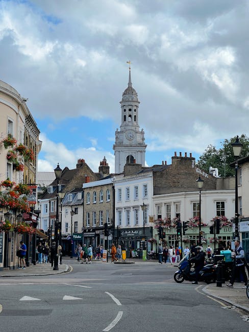The image depicts a lively street scene in Forest Gate with a prominent historic clock tower in the background under a partly cloudy sky. The foreground shows a busy intersection with pedestrians cros
