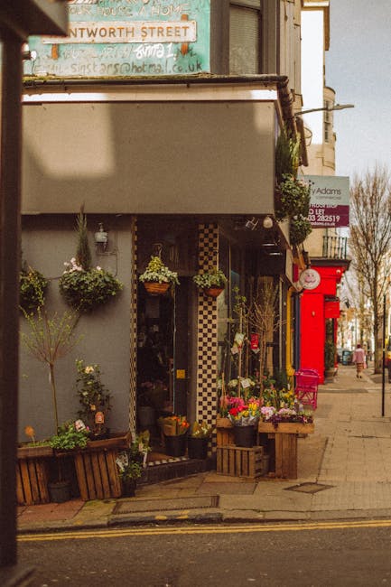 A storefront florist displaying a variety of fresh flowers arranged in baskets and pots outside on the sidewalk, including pink, purple, and white blooms, with lush green foliage. The flowers are posi
