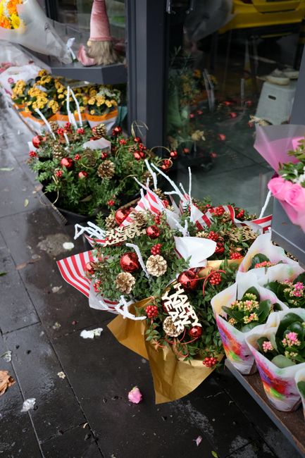 A festive floral arrangement featuring a lush bouquet of evergreen branches, pinecones, and red and white berries, accented with shiny red baubles and a small 'Valentine's Day' sign. The bouquet is wr
