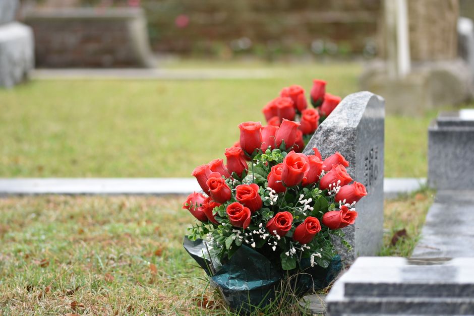 A bouquet of fresh, vibrant red roses arranged with small white filler flowers and green foliage, wrapped in dark green glossy paper. The bouquet is placed against a grey stone gravestone at a cemeter