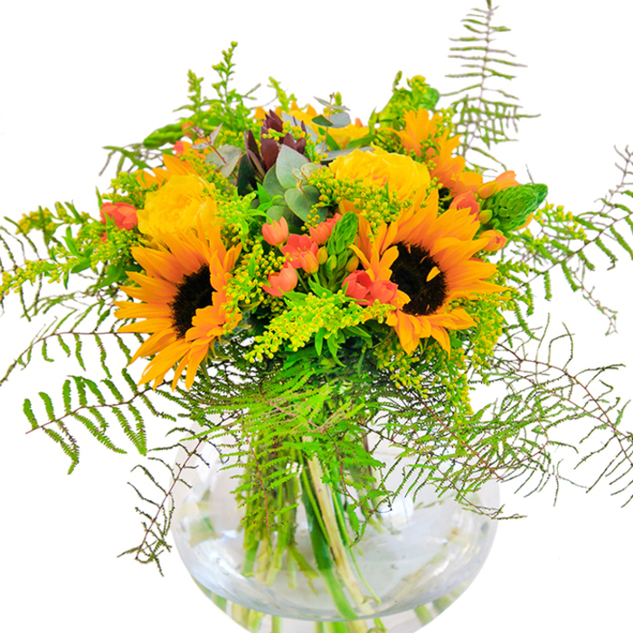 Upon first glance, this radiant bouquet crafted in Forest Gate is a portrait of seasonal exuberance-sunflowers and yellow garden roses intertwine seamlessly, their petals beaming with luminous shades of gold and amber. The contrast between the suede-like texture of the sunflowers and the tightly layered, rose-shaped blooms adds a touch of elegance that recalls the blend of old and new along Forest Gate's diverse streets. Among the sea of yellow, vibrant red Hypericum berries shine like polished gems, thoughtfully nestled beside deep green foliage and delicate vines reminiscent of the wild edges of Wanstead Park. Feathery ferns softly cascade outwards, lending gentle rhythm and movement to the bouquet, as if swaying in a late summer breeze. The eye is naturally guided to the spherical glass vase, crystal-clear and modern, offering a peek into the intertwined stems submerged in water-a visual metaphor for the open, welcoming spirit of Forest Gate's neighbourhoods. A subtle blend of eucalyptus and other light green fillers with tiny yellow blossoms completes the composition, enhancing the rustic, natural feel. The scent is understated but fresh, whispering hints of sun-warmed fields just beyond Romford Road. Perfect for milestone celebrations, heartfelt thank-yous, or as a daily reminder of joy and optimism, this arrangement brings a touch of Forest Gate's vibrant community and flourishing green spaces to any occasion.